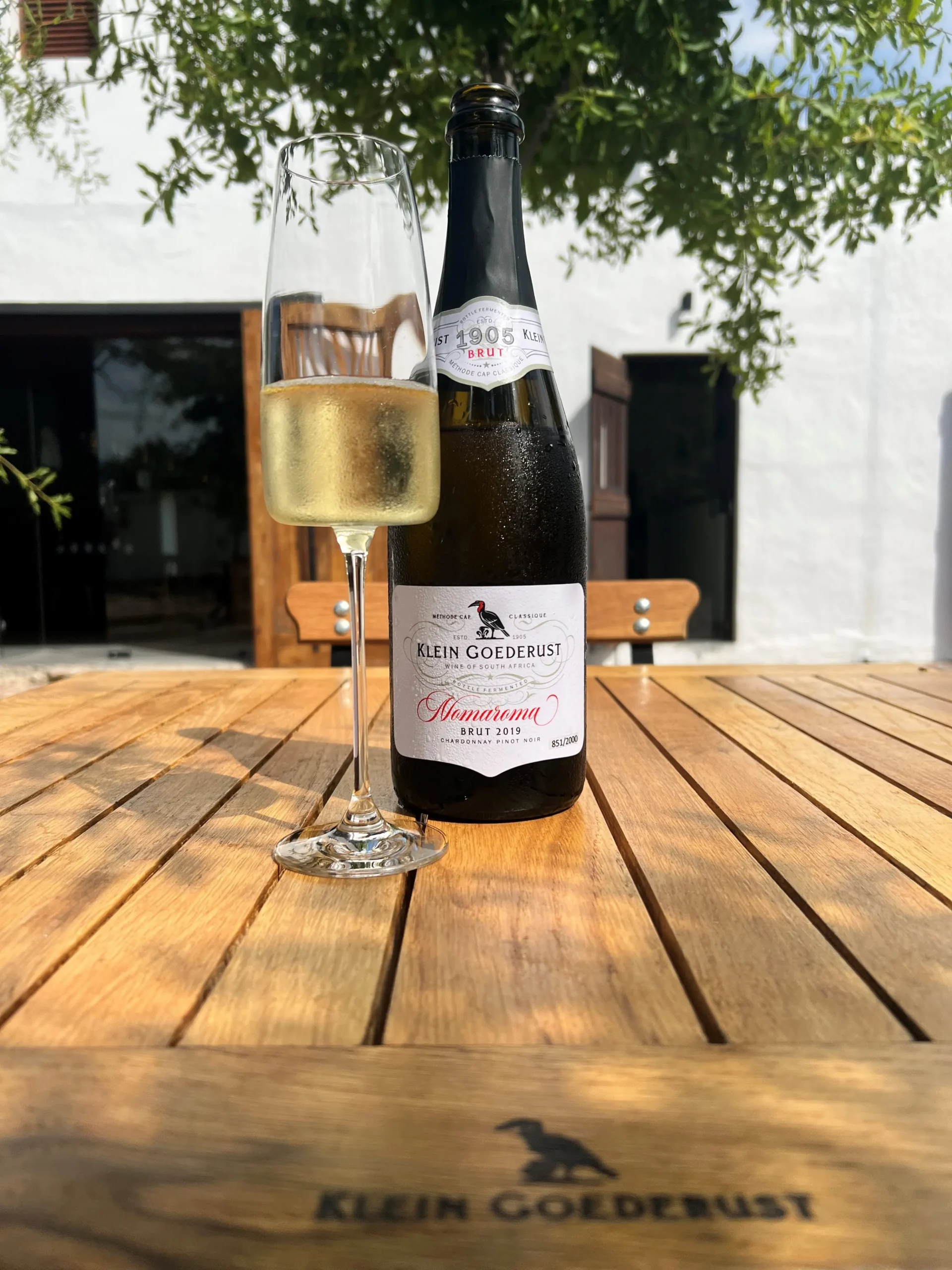 A glass of Klein Goederust Nomaroma Cap Classique beside its bottle on the estate terrace